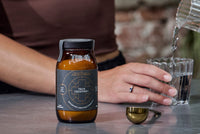 Bottle of 'True Creatine+' supplement on a table with a person pouring liquid into a glass.
