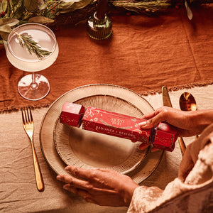 Hands holding an Ancient + Brave Radiance + Restore Cracker over a dinner plate at a table set with a cocktail glass and cutlery.