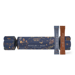An Ancient + Brave Revelry + Rest Cracker in navy blue with botanical illustrations, accompanied by two narrow, single-serving sachets in blue and brown for electrolyte replenishment.