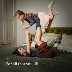 Woman lying on a bed with a child playfully lifted up on her legs in the air, with text 'For all that you lift' on a neutral background.
