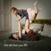 Woman lying on a bed with a child playfully lifted up on her legs in the air, with text 'For all that you lift' on a neutral background.
