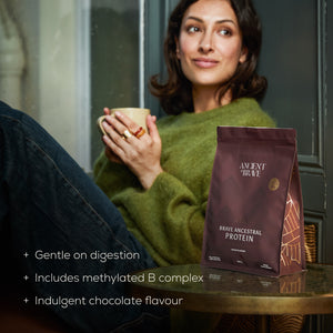 Woman holding a mug with a package of Brave Ancestral Protein on a table. With text - 'gentle on digestion, includes methylated b complex, indulgent chocolate flavour'.