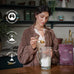 Woman preparing a protein shake with a package of Ancient + Brave's protein powder in a kitchen setting.