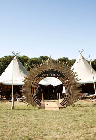 Outdoor festival scene with two large white canvas tents on a grassy field, a circular sunburst metal arch in the center, trees in the background, and several people standing around the event.
