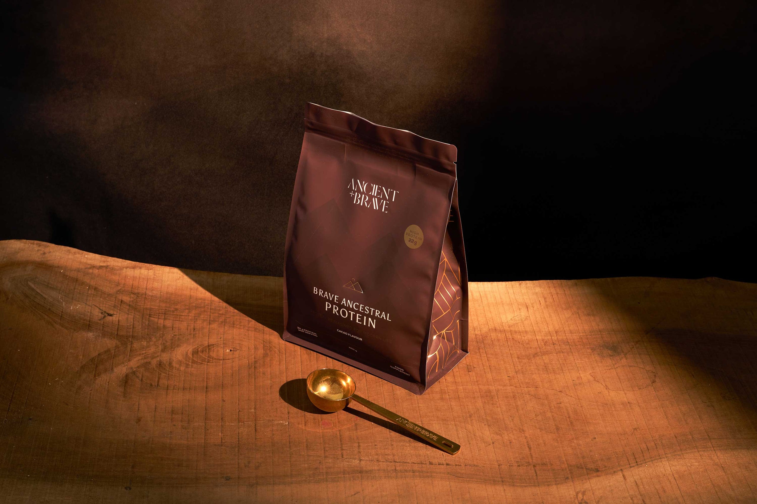 Protein powder pouch labeled 'Ancient Brewe' on a wooden surface with a gold scoop on a  dark background