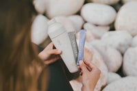 A person holds a white insulated bottle and two slim product packets in front of a blurred background of smooth white stones. The packets appear branded and include one silver packet and one blue packet.