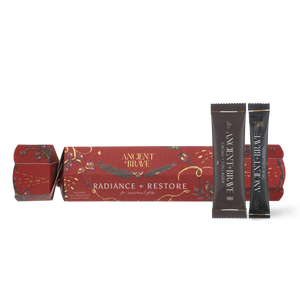 A red Ancient + Brave Radiance + Restore Cracker with gold botanical illustrations next to two single-serving sachets of cacao and collagen powder.