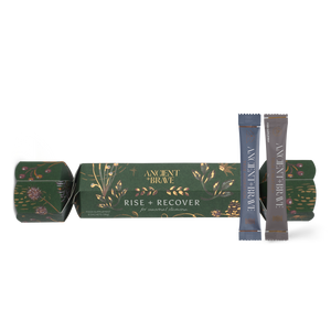 An Ancient + Brave Rise + Recover Cracker, a decorative green cardboard luxury wellbeing Christmas cracker adorned with gold botanical illustrations, accompanied by two individual creatine supplement sachets.