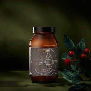 Amber glass jar of True Creatine+ Ritual Gift Set powder with a black label featuring a gold geometric cross design, positioned next to a small sprig of red berries.