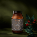 Amber glass jar of True Creatine+ Ritual Gift Set powder with a black label featuring a gold geometric cross design, positioned next to a small sprig of red berries.