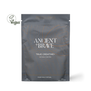 A dark gray resealable pouch labeled 'ANCIENT + BRAVE TRUE CREATINE+' with white text and thin orange line graphics, shown on a white background. A circular 'Vegan' logo appears to the left of the pouch.