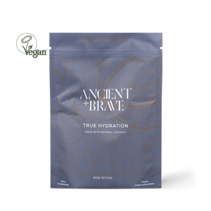 Ancient + Brawe True Hydration coconut powder supplement in a metallic slate blue pouch with gold line art, shown on a white background with a Vegan logo overlay.