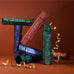 A styled arrangement of multiple Christmas crackers products in coordinated green, blue, and dusty rose packaging, with gold ribbon curls on a warm brown studio background. Visible text includes 'RISE + RECOVER,' 'REVELRY + REST,' and 'MORNING + AFTER.