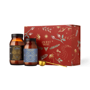 Two glass jars of collagen peptides and electrolyte powder with a gold-colored measuring scoop positioned in front of an Ancient + Brave Rise Gift Set red box decorated with gold botanical illustrations.