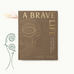 A brown, cloth-bound Ancient + Brave book titled A Brave Life with gold geometric eye line art, next to illustrations of ferns and clouds, containing rituals and recipes.