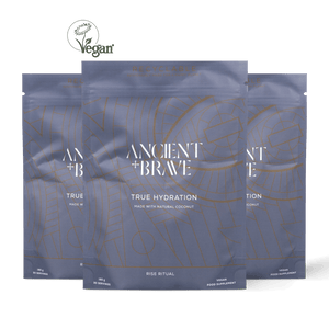 Three stand-up pouches of Ancient + Brave True Hydration food supplement arranged in a fan shape against a solid black background. The packaging is a muted slate blue-gray with white text and gold line-art patterns.