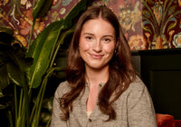 A woman with long brown hair wearing a light gray knit button-up top, smiling in front of a patterned wallpaper and a large green plant.