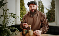 Joe Marler pouring True Biome powder from a scoop into a mug in a room with plants and a window.