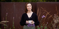A woman stands outdoors in front of a dark brown fence, holding a white mug and small purple flowers. She is wearing a black cardigan over a dark top and olive green pants.