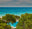 A scenic coastal view with turquoise ocean water, a sandy beach, and dense green trees in the foreground, with a small pool area visible among the foliage.