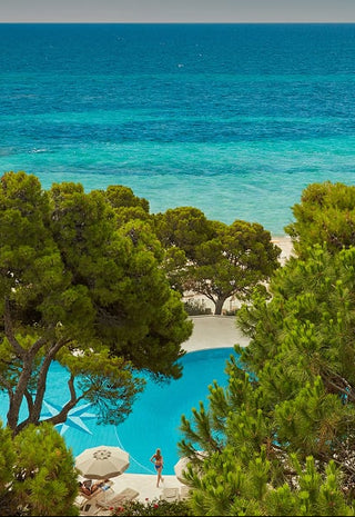 A scenic coastal view with turquoise ocean water, a sandy beach, and dense green trees in the foreground, with a small pool area visible among the foliage.