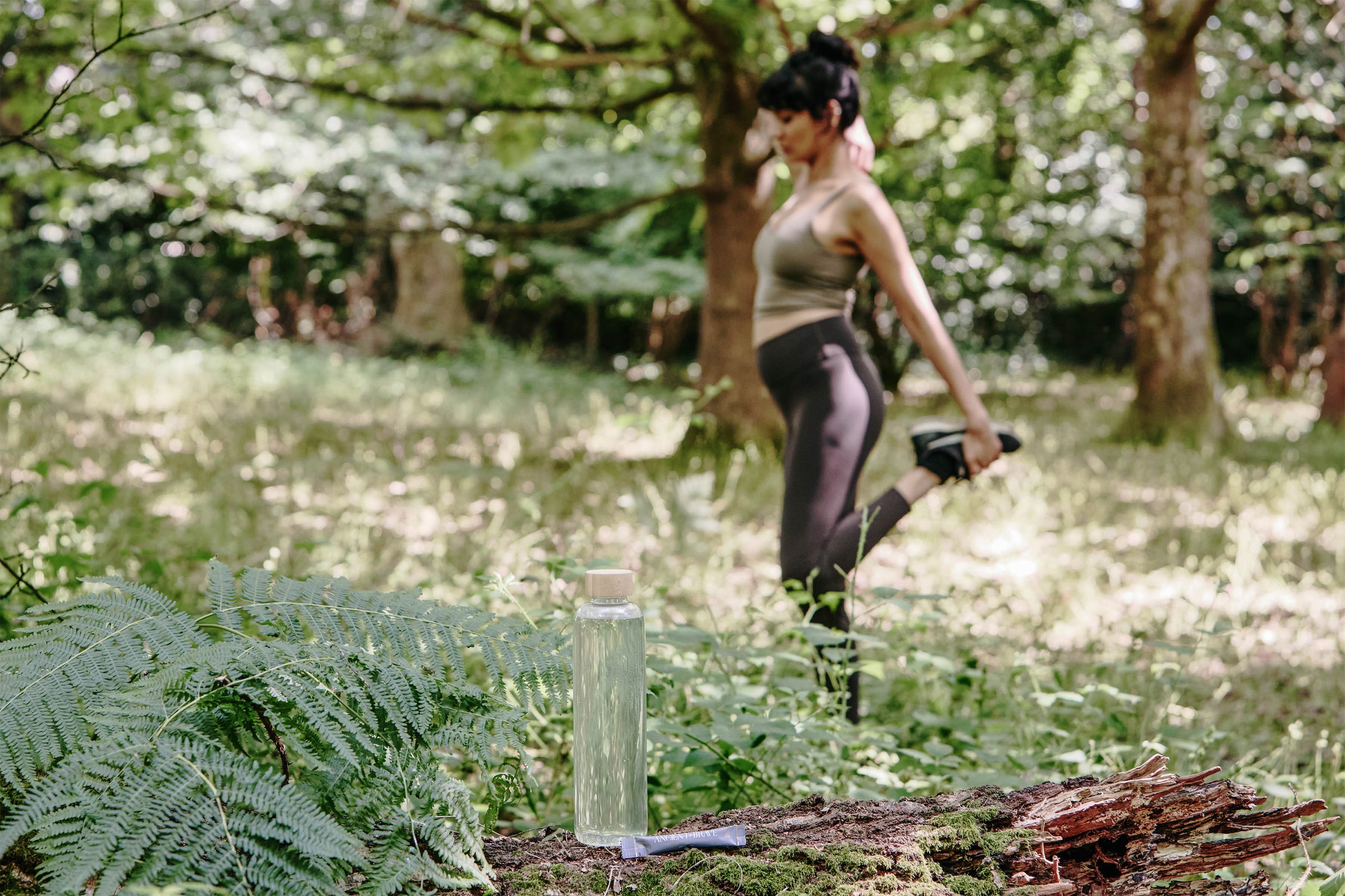 Woman stretching in a forest with a water bottle and sachet of True Hydration next to it.