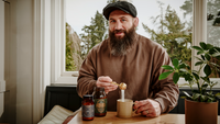 Joe Marler wearing a black flat cap and brown sweatshirt sits at a wooden table by a window, holding a small jar and spoon over a mug. Two dark glass bottles with labels are on the table, alongside a potted plant.
