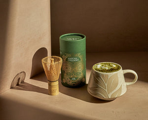A styled product shot featuring a green cylindrical canister labeled 'Matcha Collagen' from Ancient+Brave next to a bamboo whisk and a beige ceramic mug filled with matcha tea, set against a warm brown paper background.