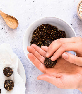 A top-down view of hands forming dark brown energy balls from a mixture, with other ingredients like oats and a wooden spoon arranged on a light gray textured surface.