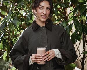 Woman holding a glass of Brave Ancestral Protein drink outdoors with greenery in the background