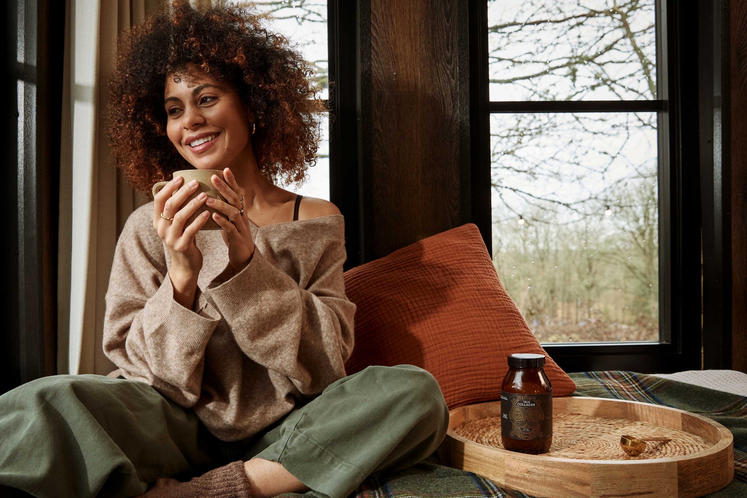 Woman sitting by a window holding a mug, with a wooden tray and bottle on a table in front of her.