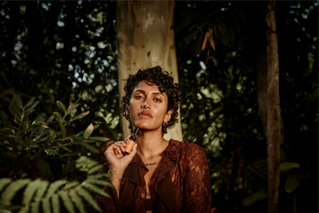 A person with short curly hair and brown lace clothing stands among the forest greenery, holding Ancient + Brave’s True Vitamin D3 + K2 - 3 Pack near their face. Sunlight highlights their features, naturally supporting vitamin D3 intake as they are surrounded by nature.