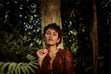 A person with short curly hair and brown lace clothing stands among the forest greenery, holding Ancient + Brave’s True Vitamin D3 + K2 - 3 Pack near their face. Sunlight highlights their features, naturally supporting vitamin D3 intake as they are surrounded by nature.