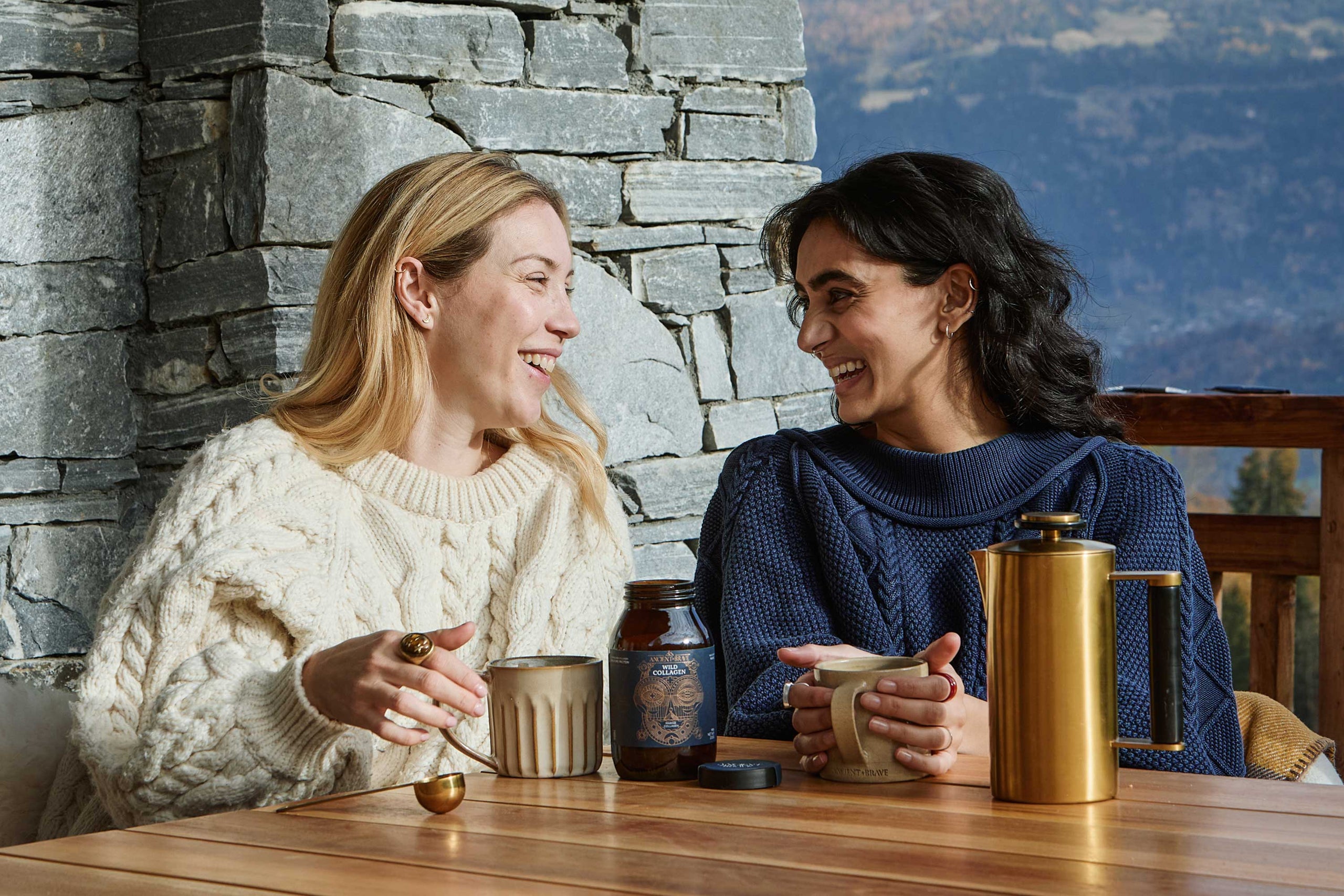 Two women sitting at a table with a mugs of coffee with Wild Collagen and cafetiere stone wall and mountain view in the background