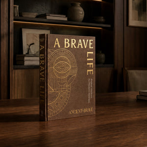 A hardcover book titled "A Brave Life" by Ancient Brave stands upright on a dark wooden table in a dimly lit room with wood paneling and shelves in the background.