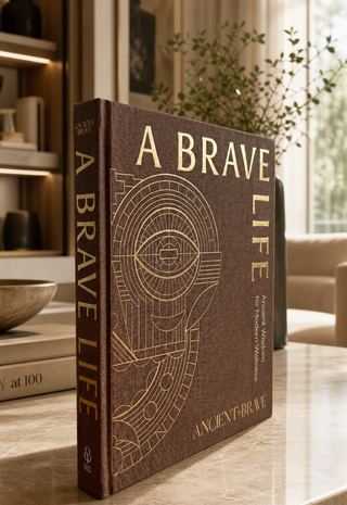 A book titled 'A BRAVE LIFE' by Ancient + Brave stands upright on a dark wooden shelf in a warm, modern interior setting with wood paneling and soft window light.