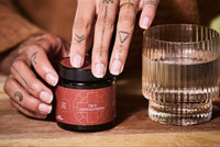 Person opening a jar labeled 'True Ashwagandha' next to a glass on a wooden surface.