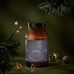 A dark amber jar of Ancient + Brave True Hydration Ritual Gift Set electrolyte powder, positioned in front of pine branches, pinecones, and a golden ribbon.