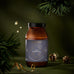 A dark amber jar of Ancient + Brave True Hydration Ritual Gift Set electrolyte powder, positioned in front of pine branches, pinecones, and a golden ribbon.