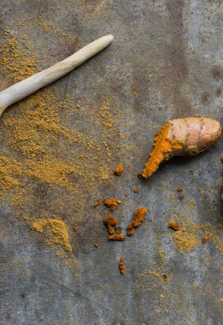 A wooden spoon with turmeric powder, fresh turmeric root, and green cardamom pods on a rustic metal surface.
