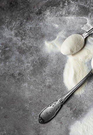 Three vintage metal spoons filled with white powder, possibly flour or sugar, rest on a dark, textured surface with some powder spilled around them.