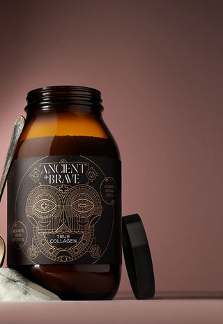 A brown glass jar labeled Ancient + Brave True Collagen sits on a surface with a metal spoon and a small heap of white powder, against a soft brown gradient background. The jar lid is lying beside it.
