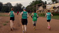 Four runners in green and blue vests on a dirt track with trees and buildings in the background.
