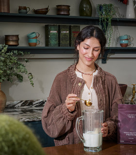 Woman pouring scoop of Ancient + Brave protein powder into milk in glass pitcher in a kitchen setting