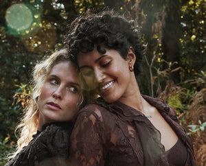 Two women embracing outdoors in a forest setting, with one wearing a dark brown lace top and the other in a black ruffled top.