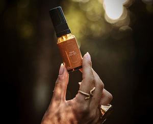A hand holding a small amber glass bottle with a black spray top and a brown label that reads TRUE D3+K2. The background is a dark, softly blurred outdoor scene with warm golden bokeh.