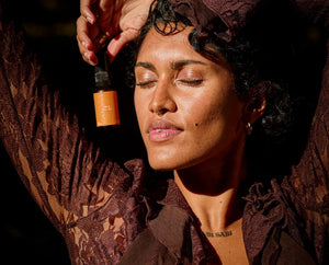 A woman with closed eyes and raised arms holds a small amber dropper bottle near her face. She wears a dark brown lace top and matching head covering in warm, dramatic lighting against a dark background.