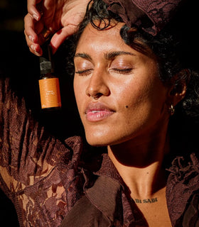 A woman with closed eyes and raised arms holds a small amber dropper bottle near her face. She wears a dark brown lace top and matching head covering in warm, dramatic lighting against a dark background.